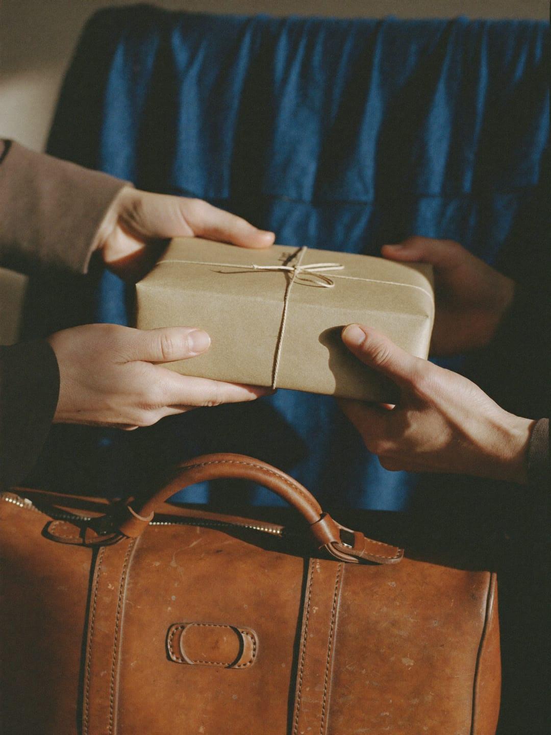 A sender handing a package to a traveler with a suitcase in a sunny park